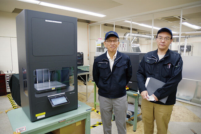 金属用3Dプリンターと泉昭一郎さん（左）、岡部経之さん