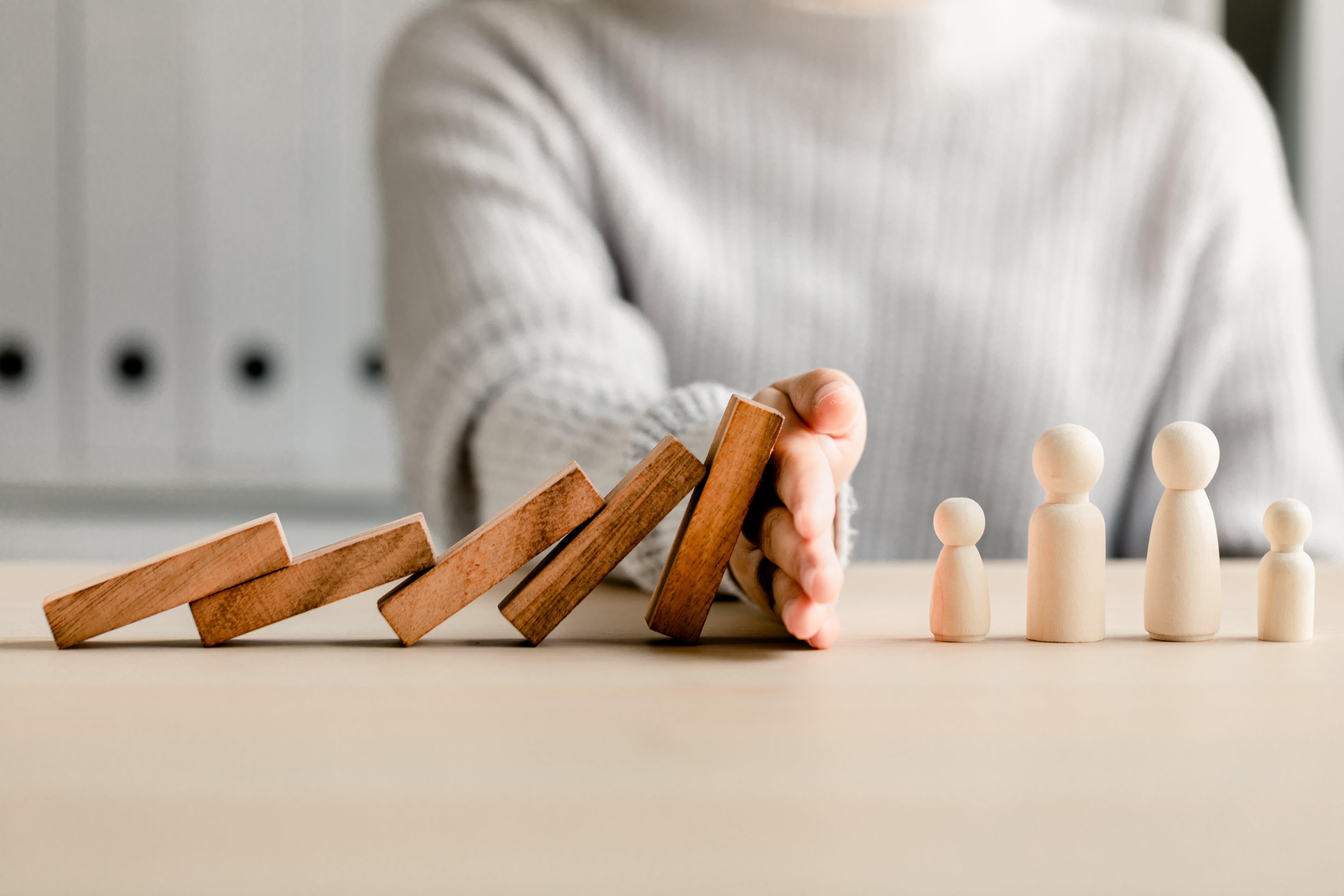 倒れかけたドミノを手で止め、後ろに並んだ人型のピースを守る様子。リスクから人や組織を守るイメージ。