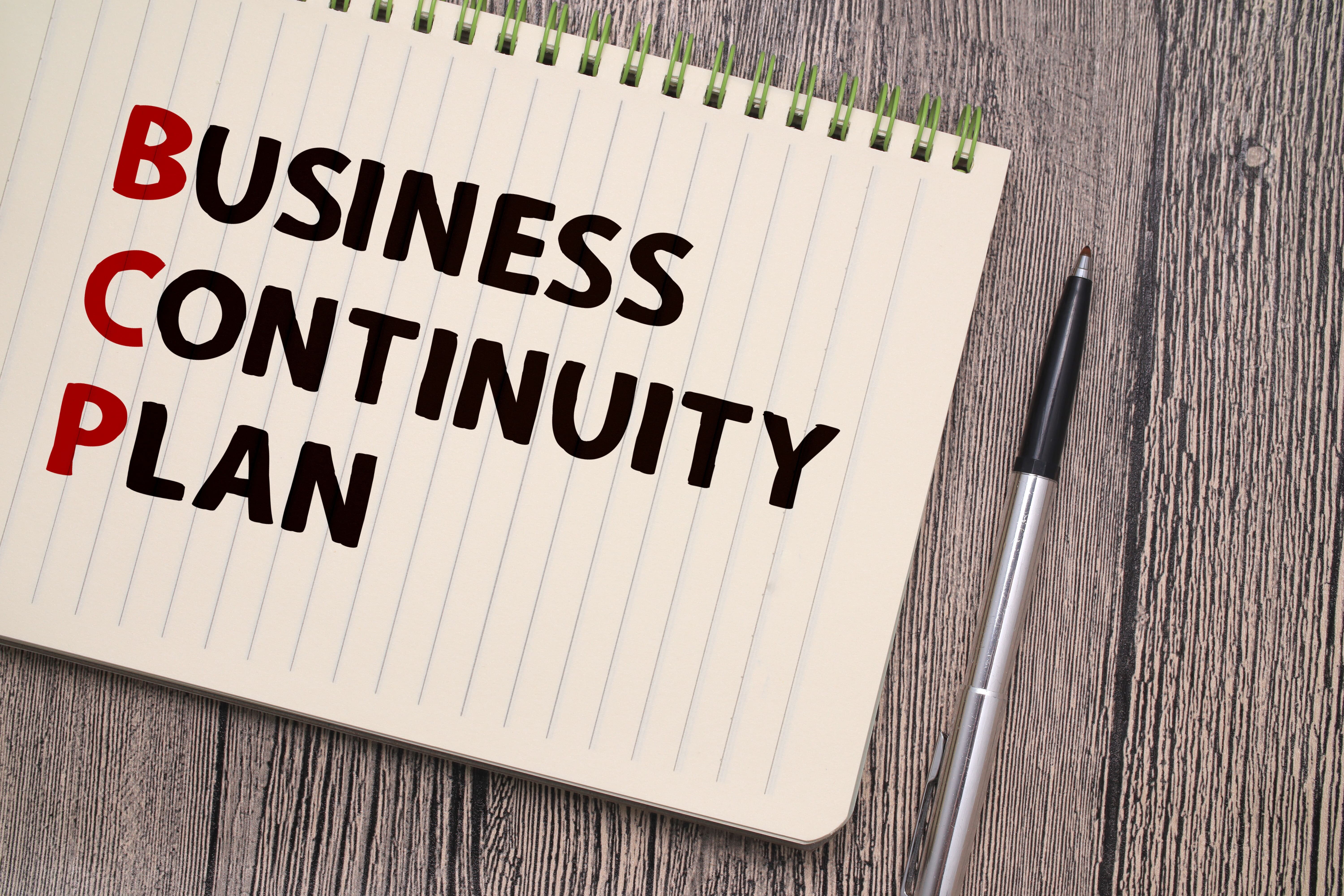 ノートに大きく“BUSINESS CONTINUITY PLAN（BCP）”と書かれた文字とペンが置かれている。BCP策定の重要性を示すイメージ。